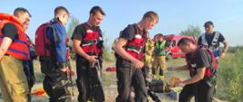 Na zdjęciu widać ratowników w kamizelkach ratowniczych. Zdjęcie w ciągu dnia. Podłoże piaszczyste. Jeden z ratowników ubiera sprzęt nurkowy.