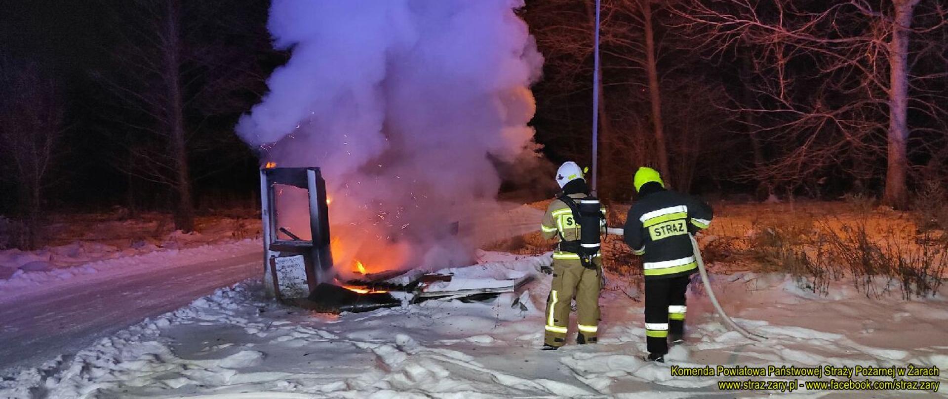 Pomimo panującej aury zimowej i grubej warstwy śniegu centralnie na zdjęciu palą się ostatnie elementy drewnianej budki parkingowej. W odległości około trzech metrów stoją dwaj strażacy i gaszą wodą palącą się budkę.