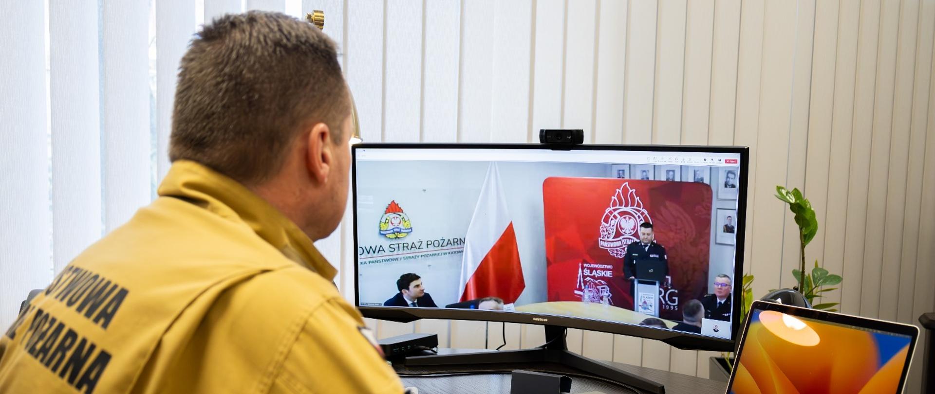 Zastępca komendanta głównego PSP w mundurze służbowym koloru piaskowego siedzi przed monitorem ekranowym podczas narady realizowanej online, w monitorze widoczny komendant wojewódzki przemawiający na mównicy podczas narady