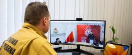 Zastępca komendanta głównego PSP w mundurze służbowym koloru piaskowego siedzi przed monitorem ekranowym podczas narady realizowanej online, w monitorze widoczny komendant wojewódzki przemawiający na mównicy podczas narady