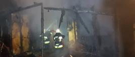 Część domku letniskowego całkowicie spalona w pożarze. W środku dwóch strażaków którzy dogaszają zgliszcza. 