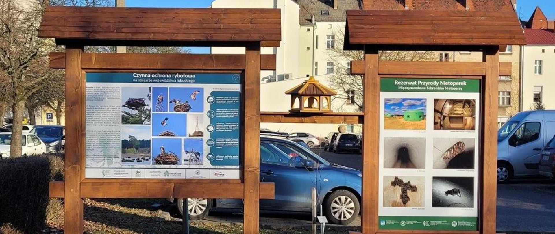 Dwie tablice informacyjne stojące na dworze. Na jednej tablicy fotografie rybołowa i opis. Na drugiej zdjęcia nietoperzy. Między tablicami stoi karmnik dla ptaków. W tle samochody na parkingu. 