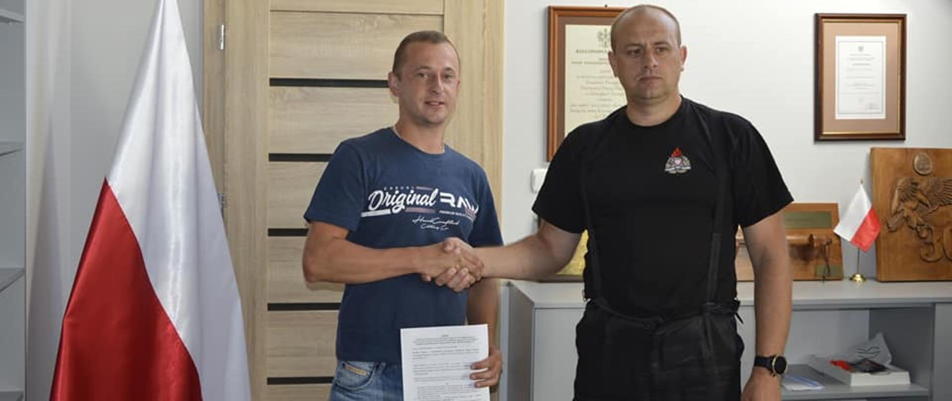 Na zdjęciu widać komendanta KP PSP w Ustrzykach Dolnych w ubraniu dowódczo-sztabowym oraz druha OSP Łodyna którzy uściskają sobie dłonie. Strażak OSP trzyma w ręku umowę. Znajdują się Oni w biurze komendanta gdzie po lewej jest flaga państwowa, po prawej dyplomy mała flaga oraz zawieszone na ścianie godło Polski.