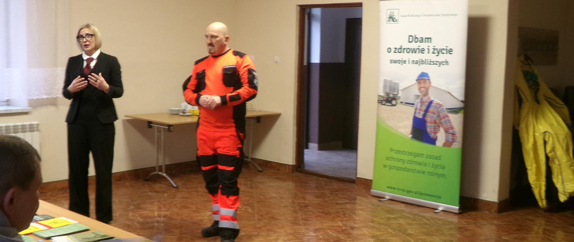 Osoby stoją na tle roll-upów KRUS, przed mini pry stole siedzą rolnicy, którzy słuchają wygłaszanej prelekcji. 