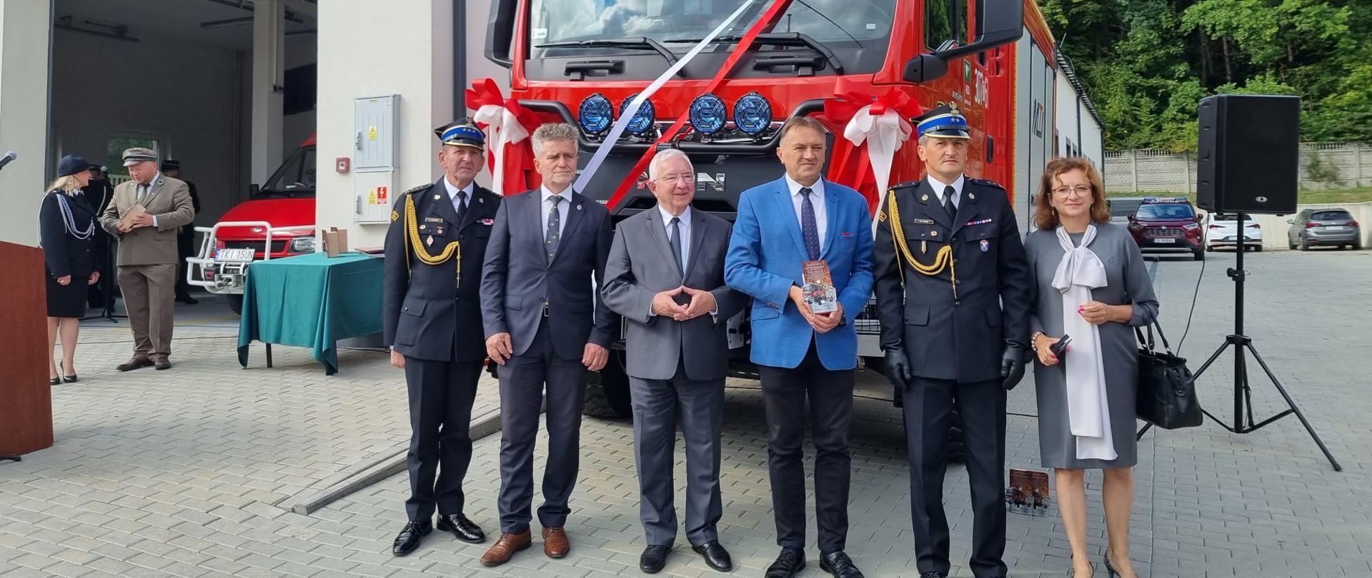 Zdjęcie przedstawia nowy samochód pożarniczy jednostki Ochotniczej Straży Pożarnej w Szewcach na którego tle stoją zaproszeni goście.