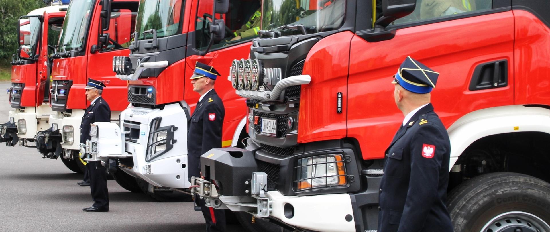 Parking przed Komendą Miejską PSP w Wałbrzychu. Na nim stoją cztery czerwone wozy strażackie, miedzy nimi stroją strażacy w żółtych mundurach bojowych