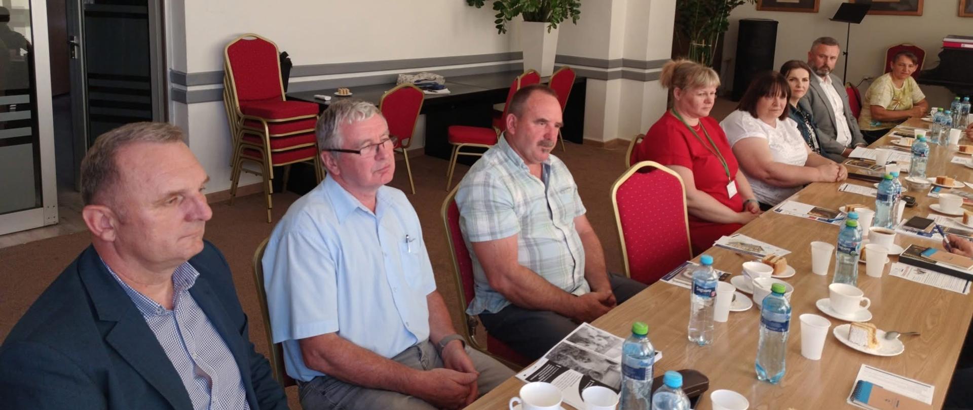 Grupa ludzi siedząca przy stole. Na stole leży woda, białe filiżanki i dokumenty. Pomieszczenie jest jasne. W tle widać zielone kwiaty i czerwone krzesła. 