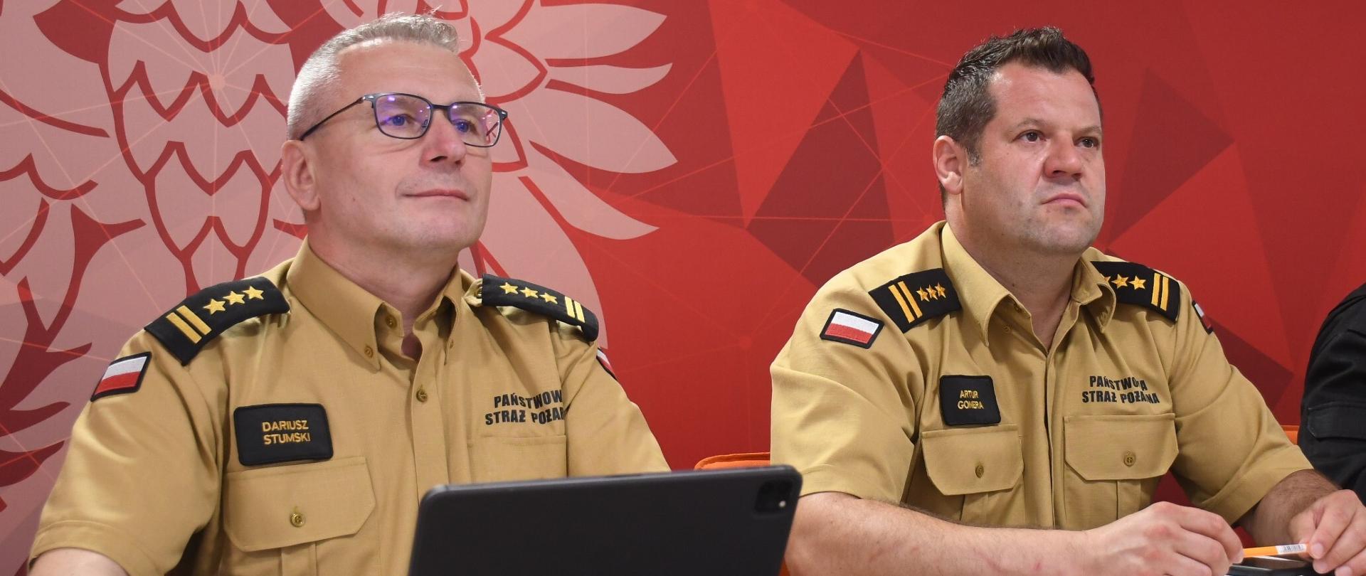 Zastępcy Mazowieckiego Komendanta Wojewódzkiego Państwowej Straży Pożarnej siedzą przy biurku. Na biurku znajduje się tablet, klawiatura i dokumenty. W tle czerwona ścianka Państwowej Straży Pożarnej.