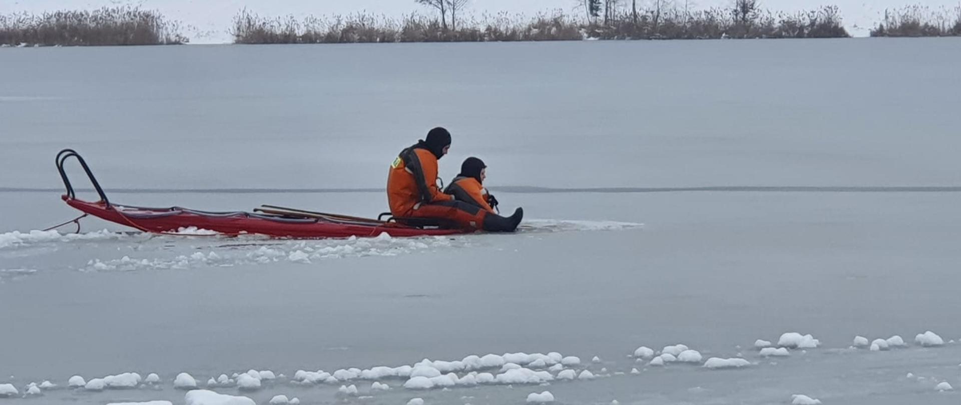 Zdjęcie przedstawia dwóch strażaków na desce lodowej na zamarzniętym akwenie wodnym 