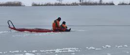 Zdjęcie przedstawia dwóch strażaków na desce lodowej na zamarzniętym akwenie wodnym 
