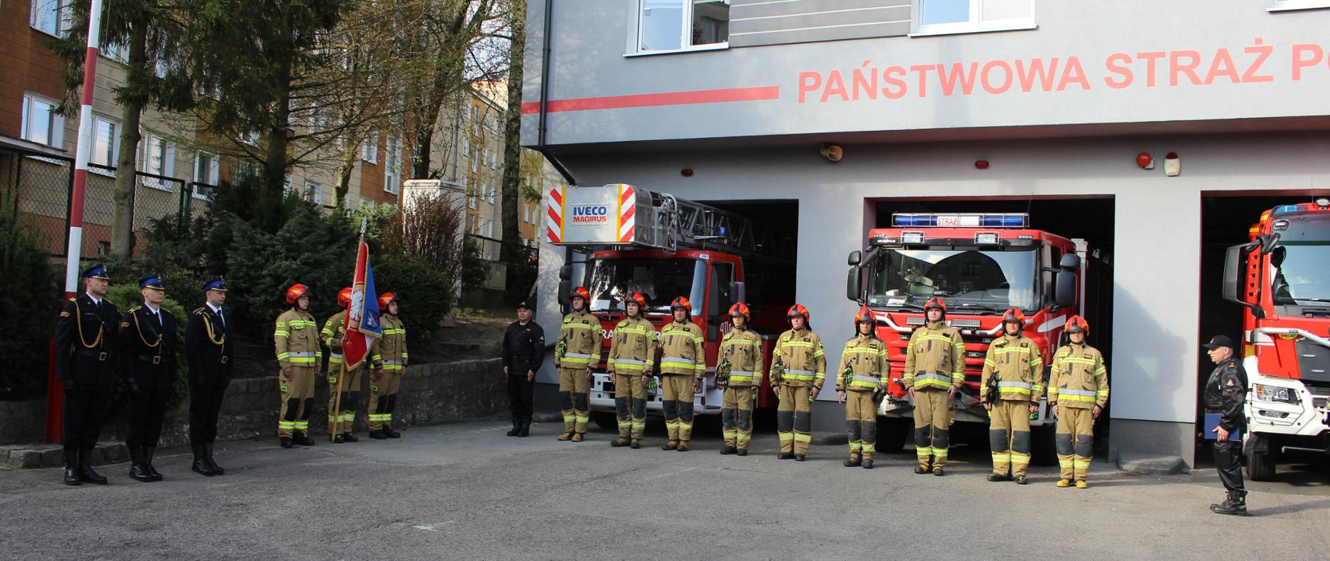 Strażacy w mundurach i hełmach stoją przed budynkiem przed uroczystym wciągnięciem flagi na maszt.