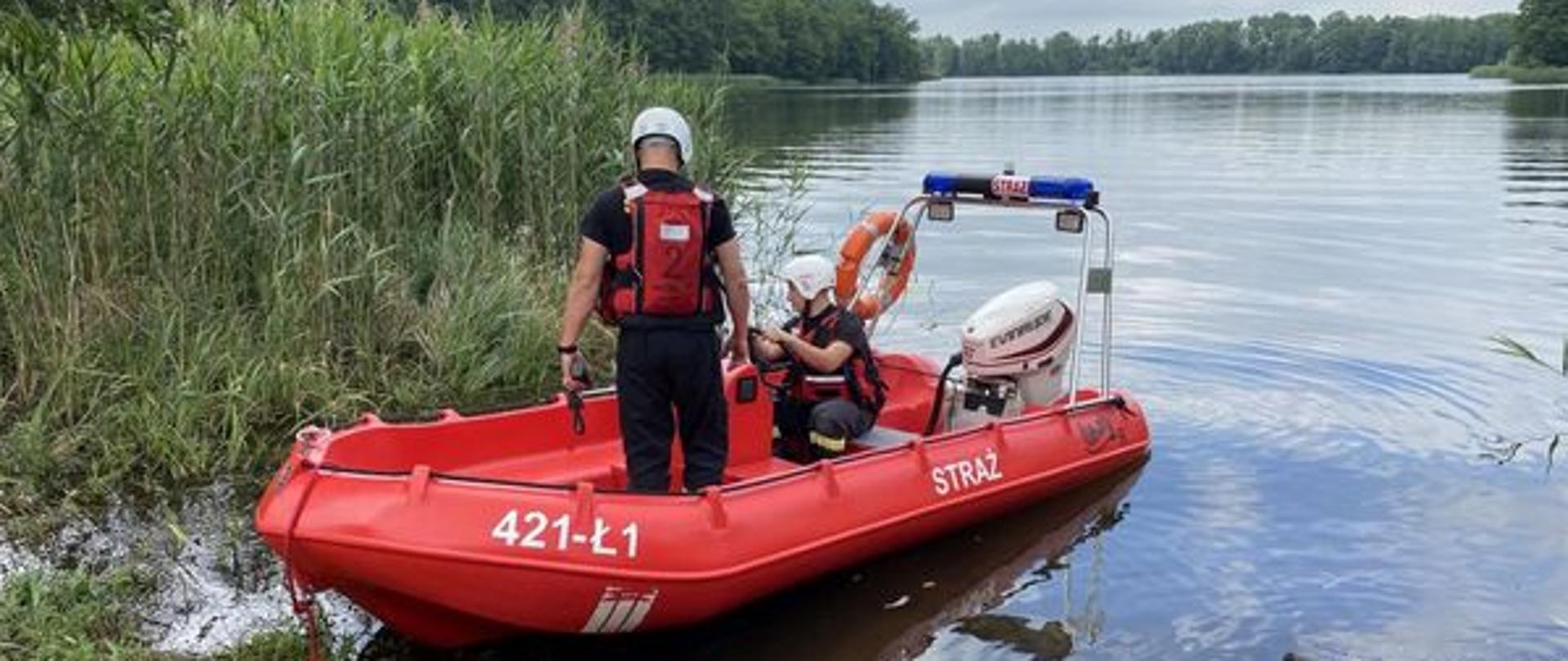 Strażacy ubrani w białe kaski do ratownictwa wodnego i kamizelki asekuracyjne na zwodowanej łodzi motorowej. Pochmurne niebo, spokojna tafla jeziora, odbijająca rosnące przy jeziorze wysokie drzewa. Za łodzią wysoka trzcina.