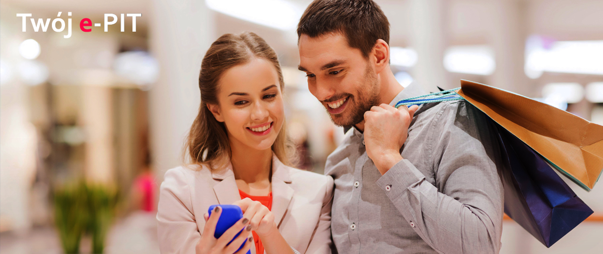 Mężczyzna trzymający torbę i kobieta patrzą uśmiechnięci na telefon komórkowy na tle galerii handlowej. W górnym lewym rogu napis "Twój e-PIT"