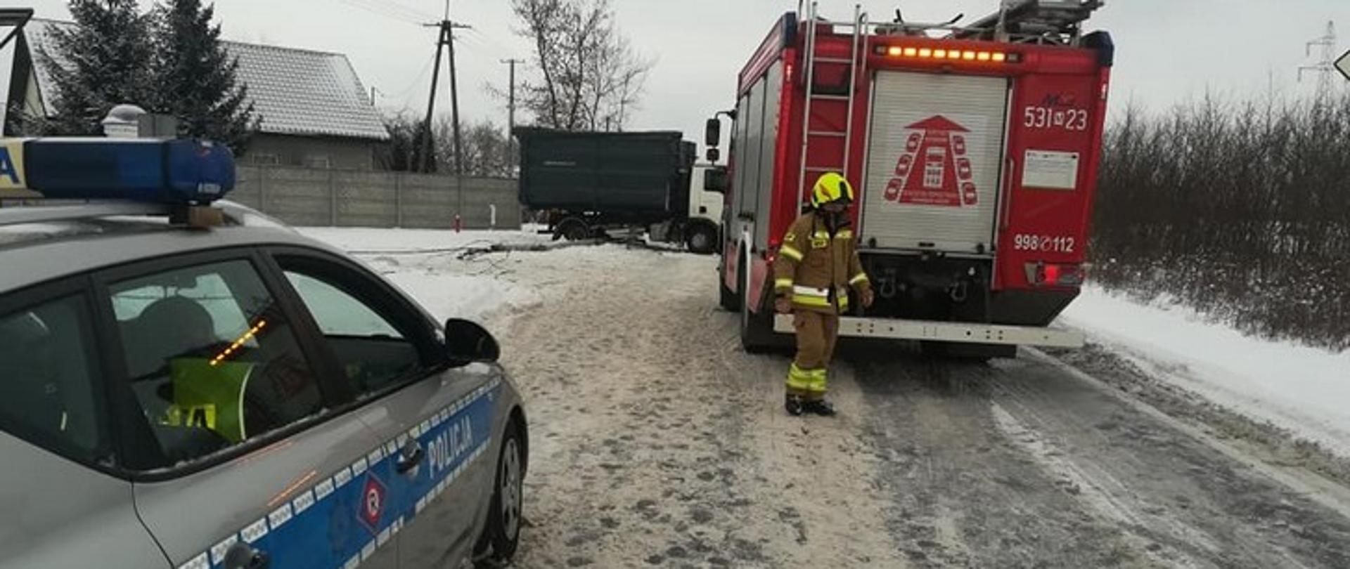 Na zdjęciu widać samochody stojące na zaśnieżonej drodze: policyjny radiowóz oraz pożarniczy, z przechodzącym obok strażakiem. W dalszej perspektywie widać uszkodzoną ciężarówkę.