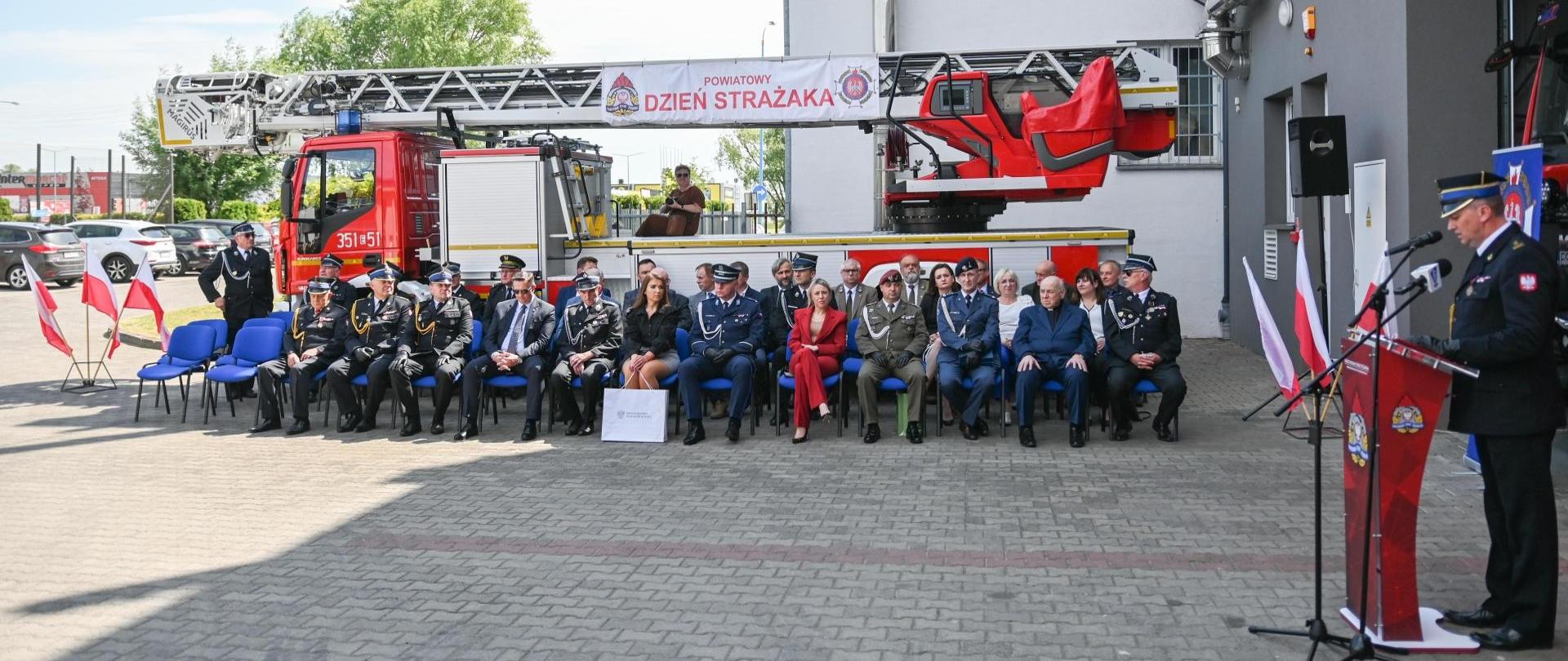 uczestnicy uroczystości siedzą na krzesłach na tle pojazdu strażackiego w kolorze czerwonym, jeden strażak przemawia.