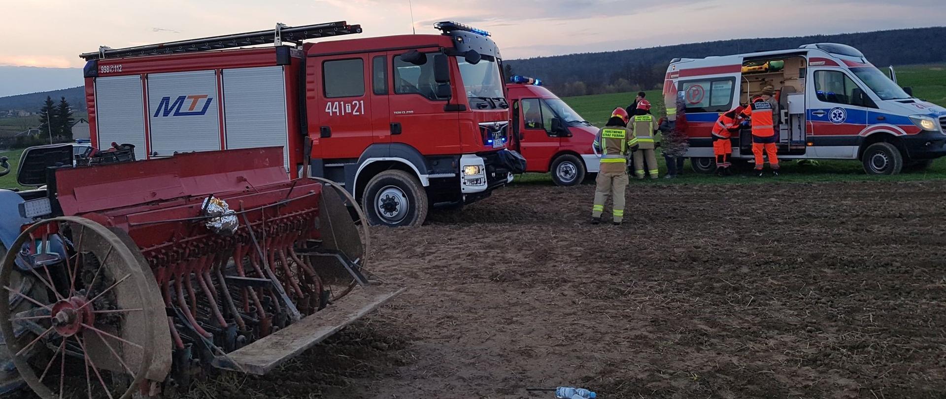 Zdjęcie przedstawia siewnik rolniczy przypięty do ciągnika rolniczego, dwa czerwone samochody pożarnicze oraz samochód Zespołu Ratownictwa Medycznego. Przed pojazdami służb ratowniczych znajdują się funkcjonariusze Straży Pożarnej, Policji oraz ratownicy medyczni