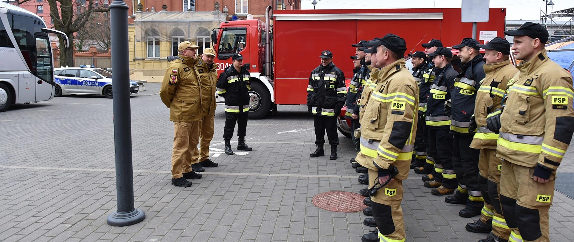 strażacy stojący na zbiórce w dwuszeregu, na przeciwko nich dwóch komendantów w tle samochód strażacki i budynki