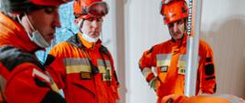 Strażacy ubrani w pomarańczowe kombinezony na głowie mają ubrane czerwone kaski, ustawiają podporę w pomieszczeniu. W tle widoczne okno.