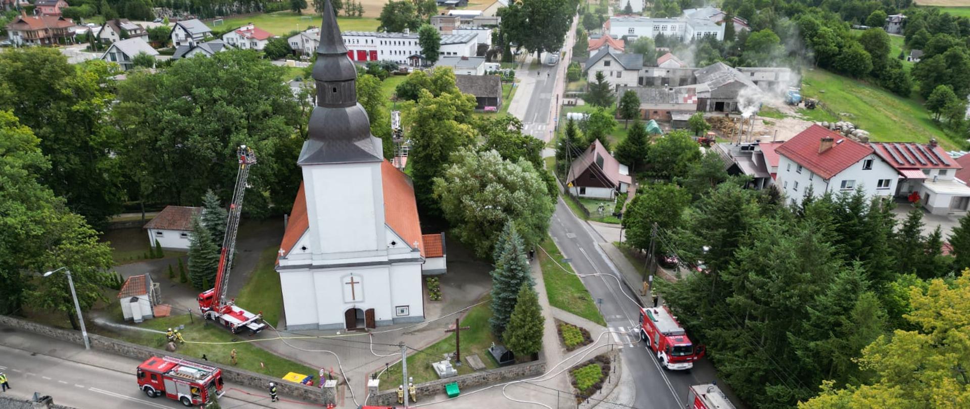Widok z lotu ptaka. Kościół na którym odbywają się ćwiczenia. Wokół kościoła na ulicy zgromadzone wozy strażackie uczestniczące w ćwiczeniach. Policja blokuje ruch. na czas ćwiczeń. Obok kościoła podnośnik z rozwiniętą drabiną. W koszy podnośnika znajdują się strażacy. W tle zabudowania wsi oraz pola i lasy.