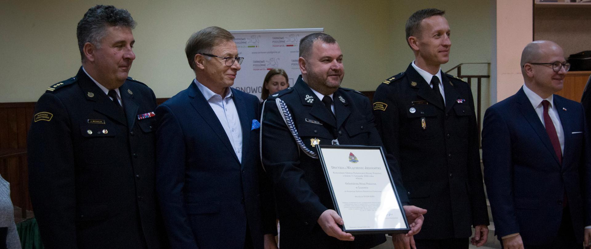 trzech strażaków oraz dwoch mężczyzn stoi obok siebie. strażak w środku trzyma ramkę z aktem powolania