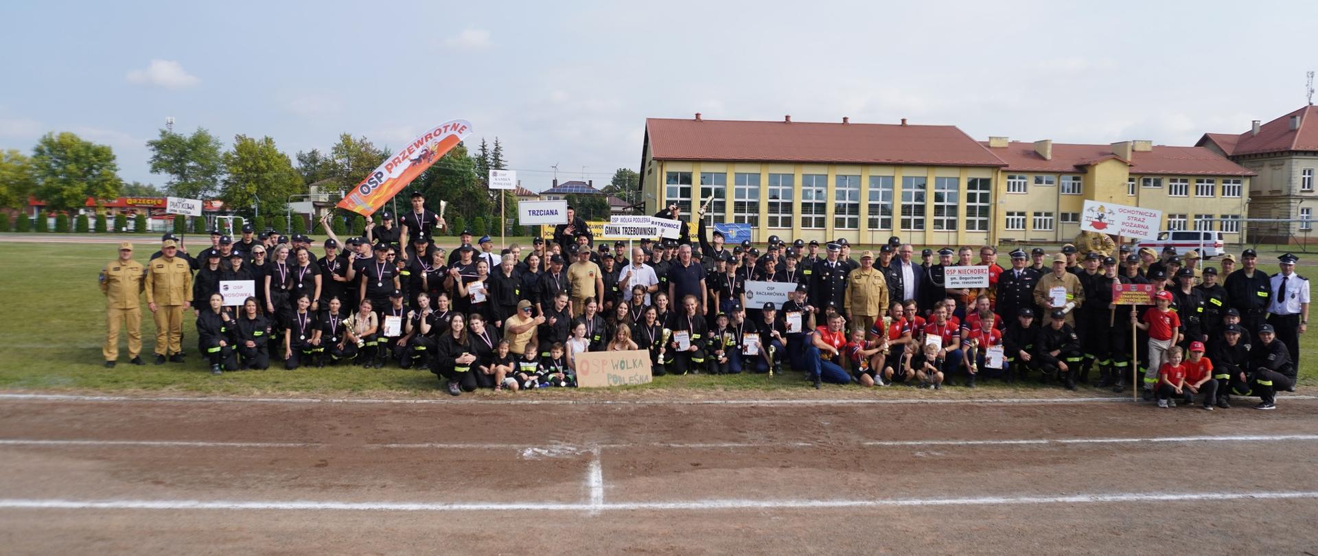 Duża grupa uczestników zawodów strażackich, w tym dzieci, młodzież i dorośli w różnych mundurach i strojach drużynowych, pozuje wspólnie do pamiątkowego zdjęcia na boisku. Widoczne są transparenty z nazwami jednostek, medale i dyplomy. W tle znajduje się budynek szkoły.