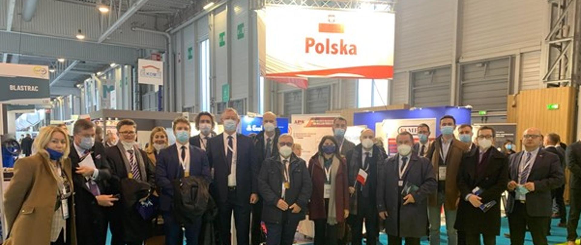 Zdjęcie przedstawiające grupę ludzi stojących przed stoiskiem na targach branży jądrowej World Nuclear Exhibition 2021, w tle nad głowami ludzi widoczny jest napis Polska z polską flagą ponad nim 