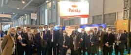Zdjęcie przedstawiające grupę ludzi stojących przed stoiskiem na targach branży jądrowej World Nuclear Exhibition 2021, w tle nad głowami ludzi widoczny jest napis Polska z polską flagą ponad nim 