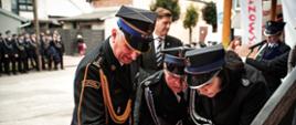 Strażacy w mundurach galowych uczestniczą w ceremonii wbijania pamiątkowych gwoździ w drewnianą tablicę sztandarową lub pamiątkową. Używają do tego specjalnych młoteczków, skupiając uwagę na precyzyjnym wykonaniu gestu.