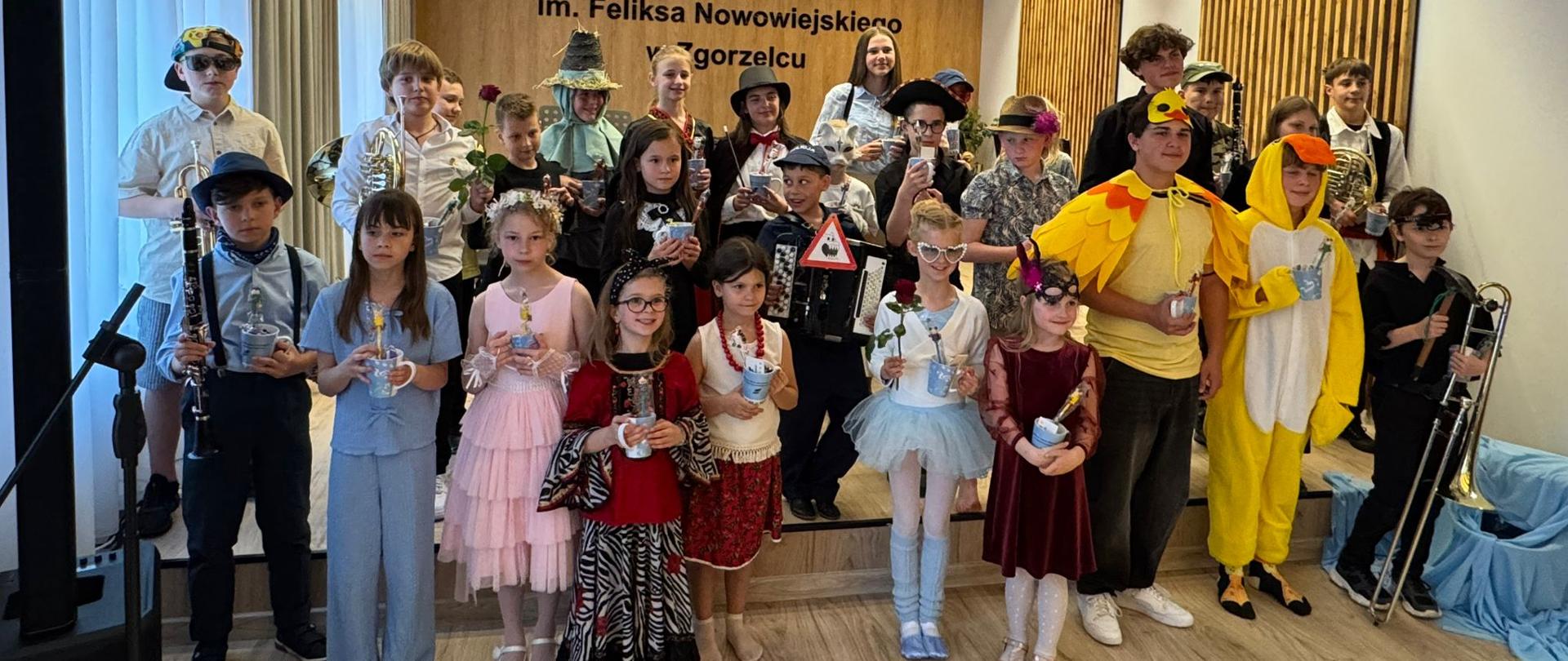 Zdjęcie grupowe uczestników koncertu Figle muzyczne, dzieci w kolorowych przebraniach stojące w trzech rzędach na scenie szkolnej auli.