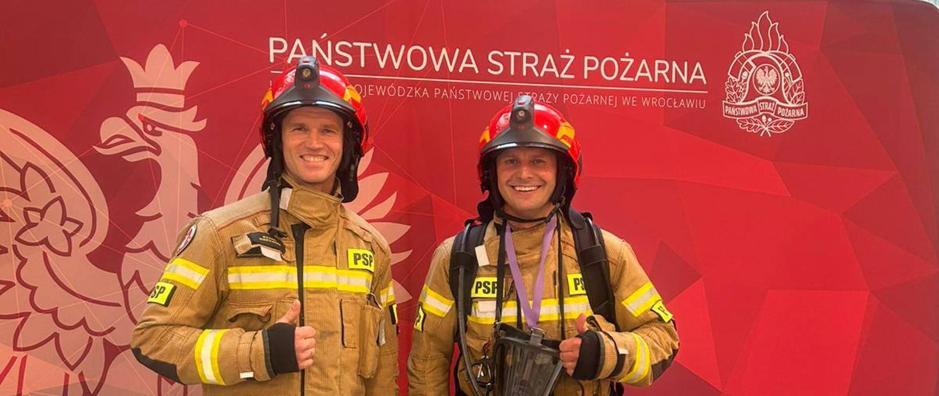 Na zdjęciu dwóch strażaków w mundurach bojowych i czerwonych hełmach stoją na tle czerwonej ścianki z napisem państwowa straż pożarna.