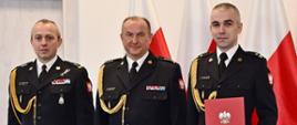 Trzech strażaków w mundurach wyjściowych ze sznurem stoi obok siebie jeden z mężczyzn trzyma czerwoną teczkę za nimi ustawione są trzy flagi Polski oraz znajdują się drzwi. 