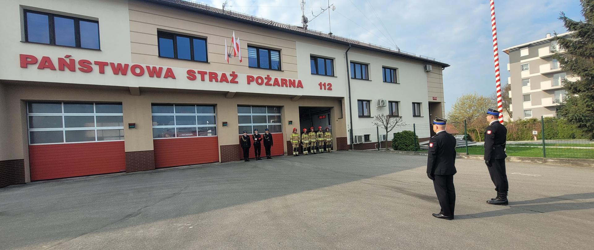 Uroczysta zbiórka z okazji Dnia Flagi Rzeczypospolitej, strażacy w ubraniach specjalnych i mundurach galowych stoją przed budynkiem komendy 