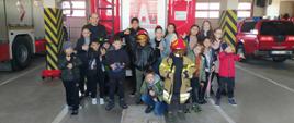 Na pierwszym planie w garażu grupka dzieci z Ukrainy pozują do fotografii, unoszą do góry prawy kciuk, jeden chłopiec na środku kadru ubrany jest w kurtkę bojową strażaka koloru jasnożółtego oraz ma założony hełm barwy czerwonej na głowie, w tle za nimi stoi auto ciężarowe barwy czerwonej, po lewej i prawej stronie samochody bojowe straży pożarnej