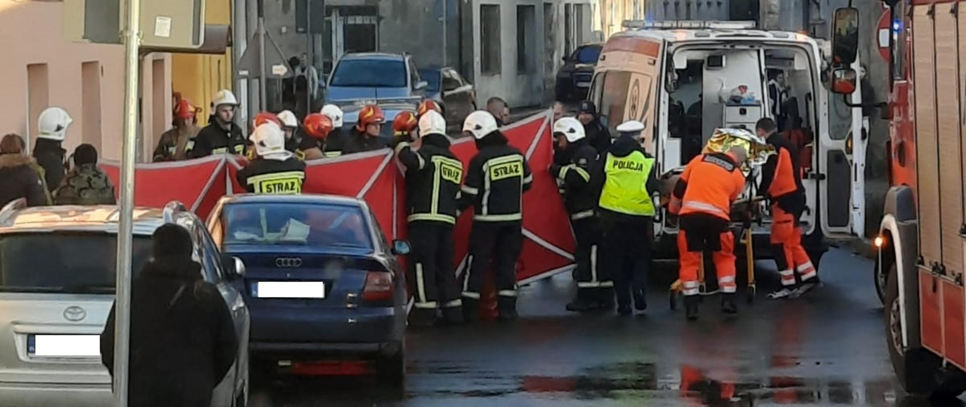 Dzień widok na ulice. W tle karetka pogotowia obok której stoi grupa strażaków w białych hełmach i ubraniach specjalnych w kolorze granatowym z żółtymi, odblaskowymi elementami. Strażacy rozstawiają parawan w kolorze czerwonym.