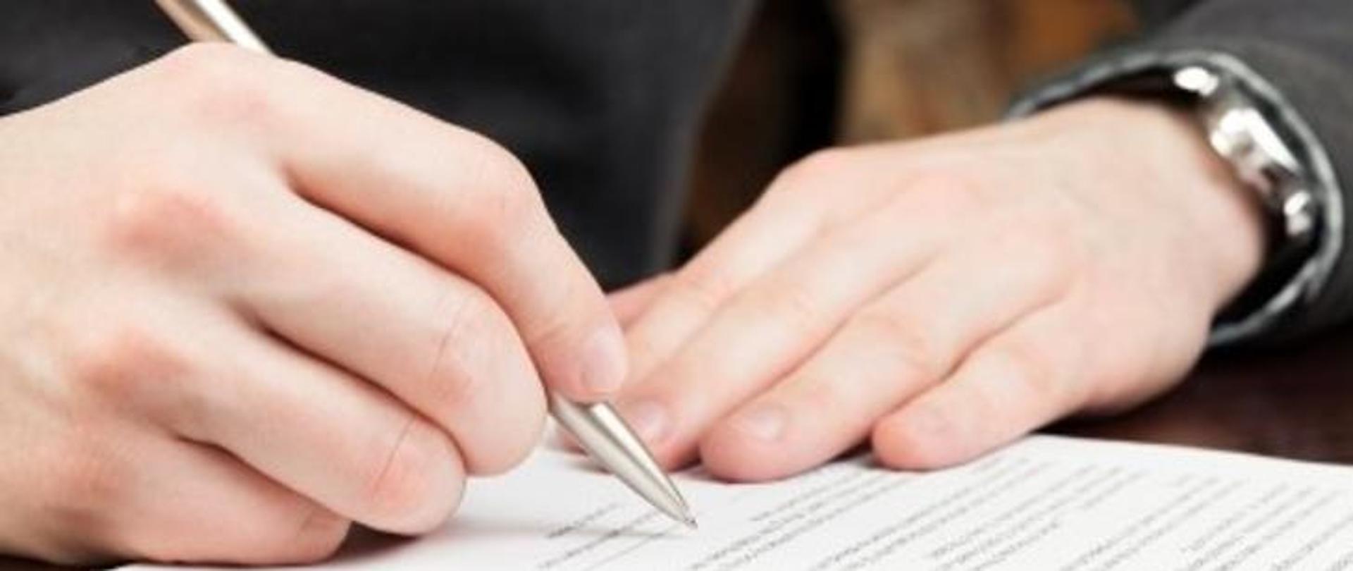 Na zdjęciu widoczny jest dokument oraz dłonie. W jednej z nich mężczyzna trzyma długopis i podpisuje dokument. 