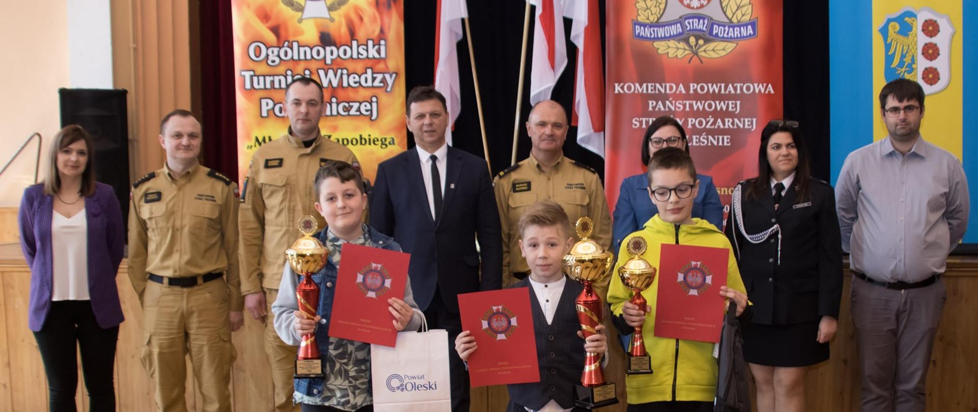 Na zdjęciu strażacy, osoby cywilne o dzieci z pucharami i dyplomami podczas Ogólnopolskiego Turnieju Wiedzy Pożarniczej.