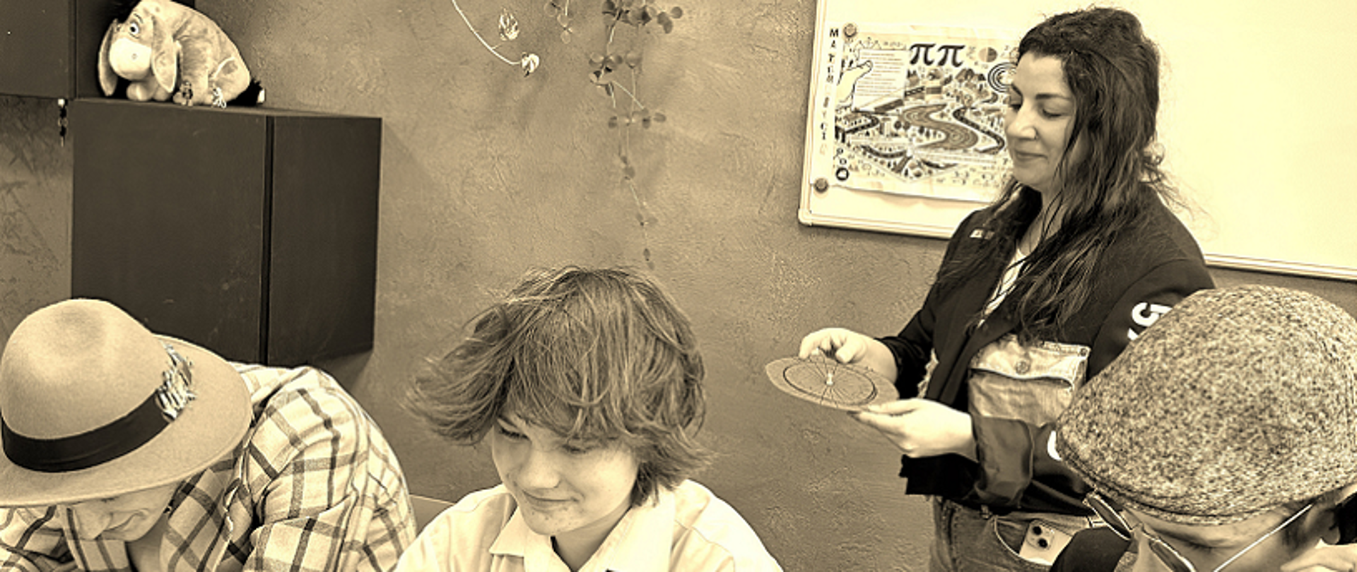 Na zdjęciu widzimy grupę uczniów zaangażowanych w pracę nad zadaniami związanymi z kryptologią. Jeden z uczniów korzysta ze starej maszyny do pisania, drugi używa lupy, a trzeci sprawdza czas na zegarku, jednocześnie dzwoniąc. W tle na tablicy widoczny jest napis „MATematycznie zakodowani” – nazwa zespołu, co podkreśla tematykę matematyczno-informatyczną ich działań. Zdjęcie utrzymane jest w stylu retro, co dodatkowo nawiązuje do historycznych metod kodowania i łamania szyfrów.