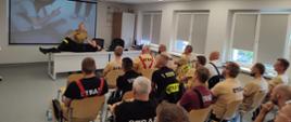 Na zdjęciu widać strażaków OSP w ubraniach koszarowych zasiadających w sali szkoleniowej Ośrodka Szkolenia KW PSP w Opolu. Z tyłu zdjęcia zajęcia prowadzi funkcjonariusz Ośrodka Szkolenia KW PSP w Opolu w umundurowaniu służbowym - demonstruje pozycję boczną bezpieczną na pozorancie. W tle wyświetlana jest prezentacja multimedialna.