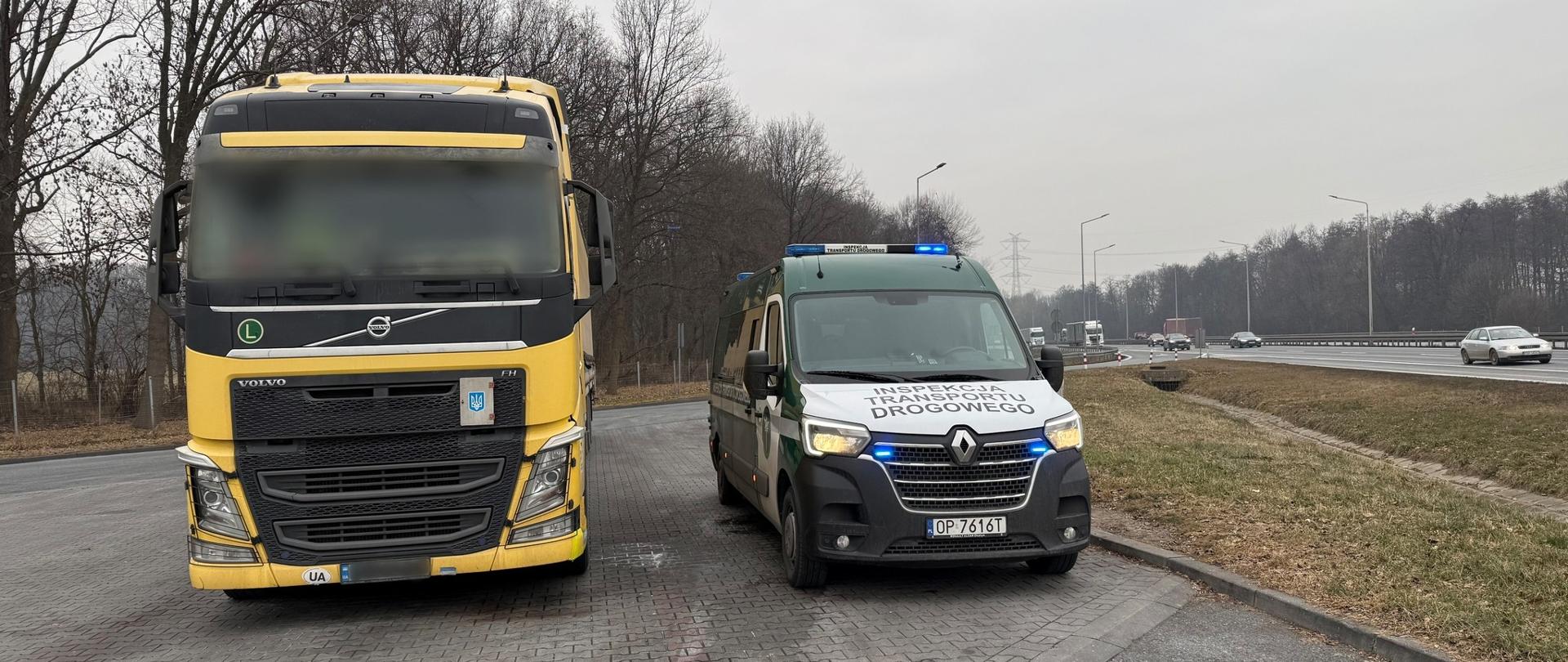 Żółta ciężarówka marki Volvo zatrzymana na poboczu drogi. Obok niej stoi pojazd Inspekcji Transportu Drogowego z włączonymi niebieskimi światłami ostrzegawczymi. Scena rozgrywa się przy drodze szybkiego ruchu, w pochmurny dzień, z widocznymi drzewami bez liści w tle.