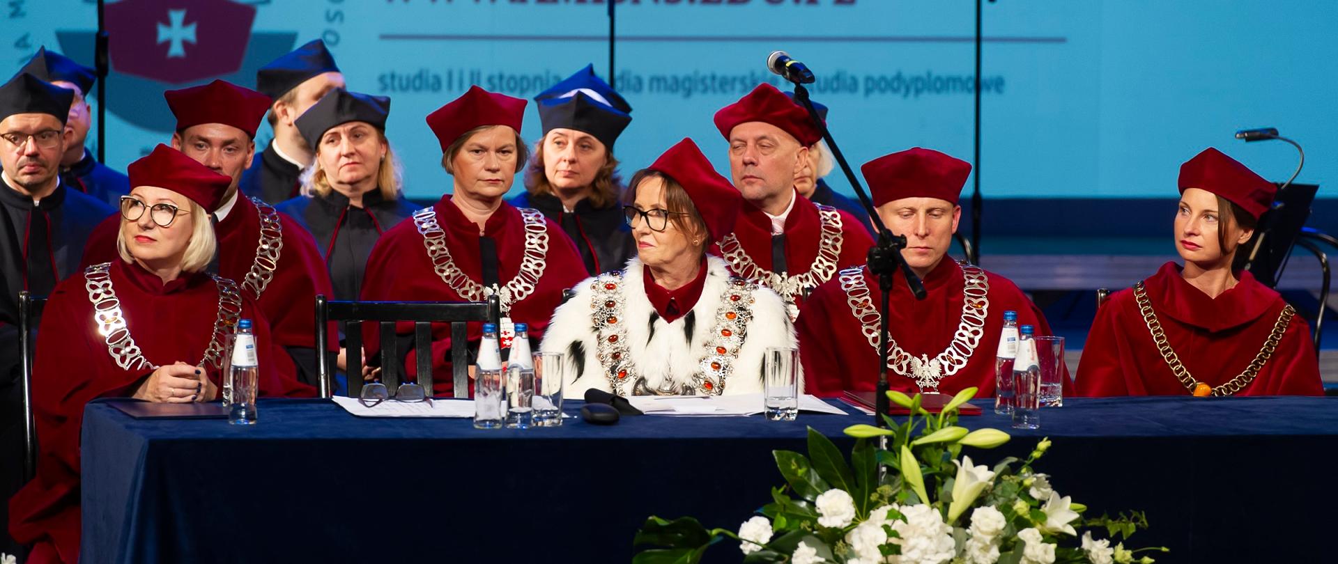 Grupa osób w czerwonych ceremonialnych strojach siedzi za niebieskim stołem, przed nim bukiet białych kwiatów.