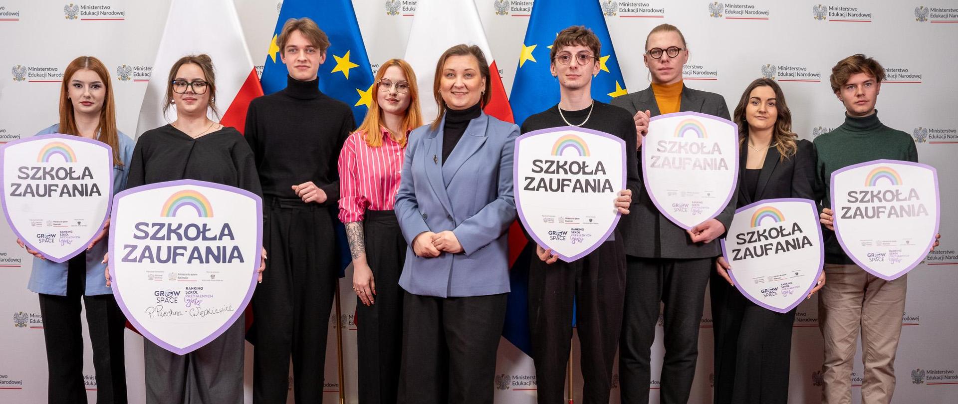 Na tle białej ścianki z napisem Ministerstwo Edukacji Narodowej stoi wiceminister Paulina Piechna-Więckiewicz, obok niej grupa młodych ludzi, którzy trzymają w rękach duże tarcze z napisem Szkoła zaufania. W tle flagi Polski i UE.