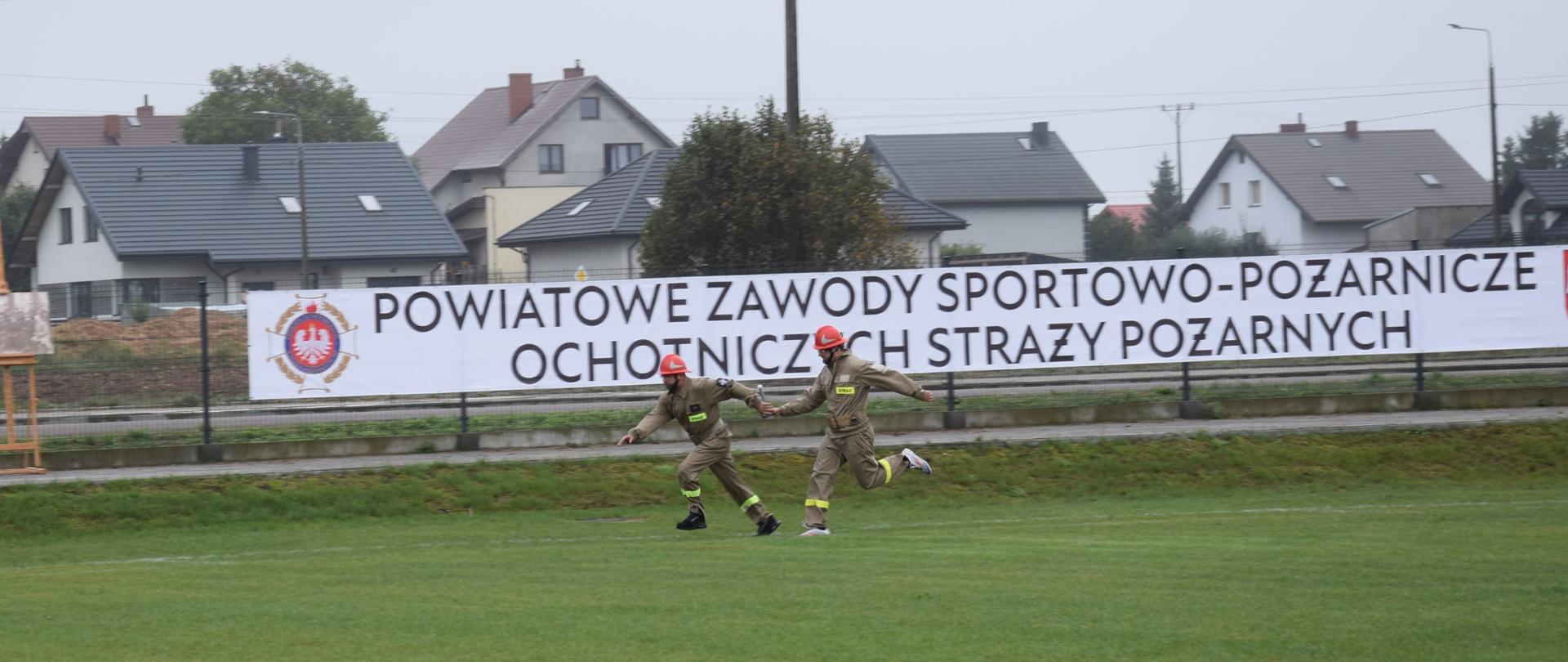 Na zdjęciu widzimy dwóch strażaków w trakcie zawodów, biegnących po trawiastym terenie. Obaj ubrani są w beżowe kombinezony i czerwone kaski. W tle znajduje się duży, biały baner z napisem "POWIATOWE ZAWODY SPORTOWO-POŻARNICZE OCHOTNICZYCH STRAŻY POŻARNYCH"