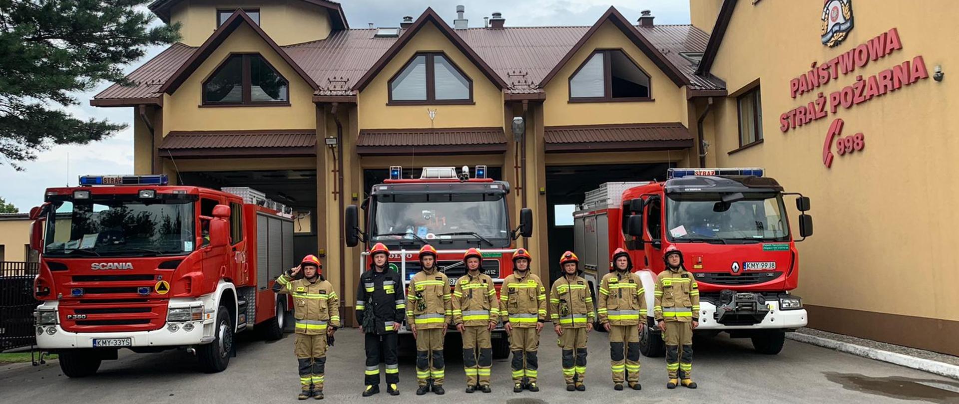 Na zdjęciu widzimy samochody strażackie przed garażami (budynkiem Komendy Powiatowej) oraz zmianę służbową stojącą przed nimi. Dowódca zmiany salutuje.