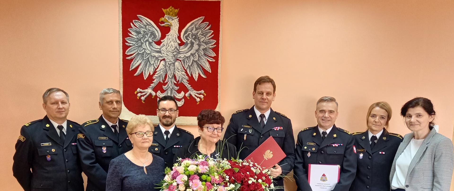 Strażacy w mundurach wyjściowych i pracownicy cywilni na tle ściany i wiszącego na niej godła RP. W środku jubilatka trzymająca bukiety kwiatów.