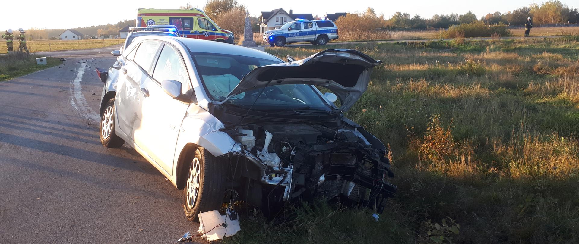 Zdjęcie przedstawia zniszczony w wyniku zderzenia samochód osobowy. Za nim w głębi stoją pojazdy służb ratowniczych.