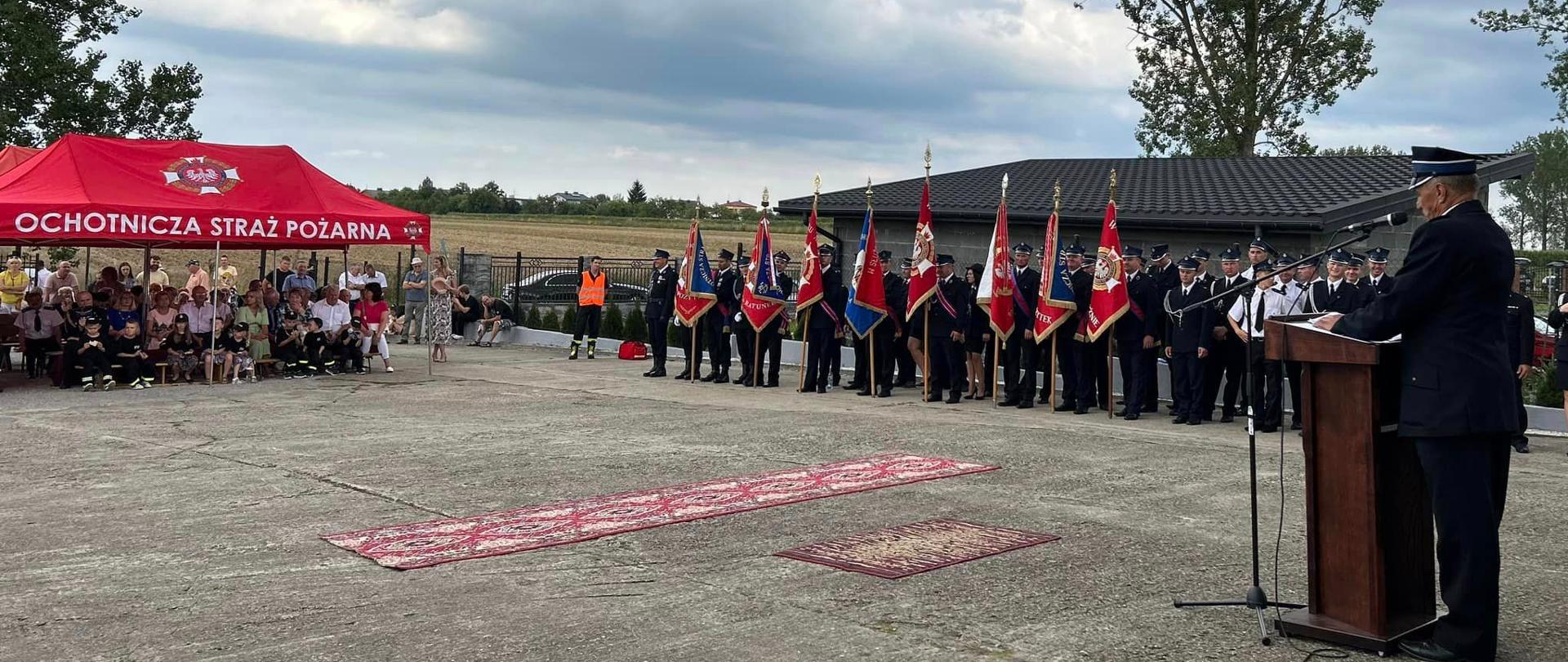 plac za jednostką osp na którym odbywa się uroczystość po prawej stoją poczty sztandarowej i druhowie w dwuszeregu po lewej na namiotem siedzą dzieci i dorośli