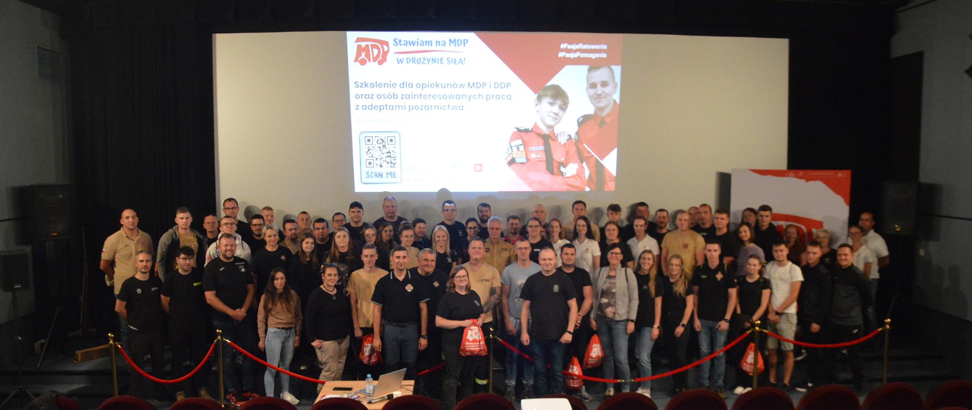 Szkolenie opiekunów MDP. Zdjęcie przedstawia salę kinową. Na tle ekranu, na którym wyświetlana jest prezentacja, stoją uczestnicy szkolenia, którzy pozują do grupowego zdjęcia.