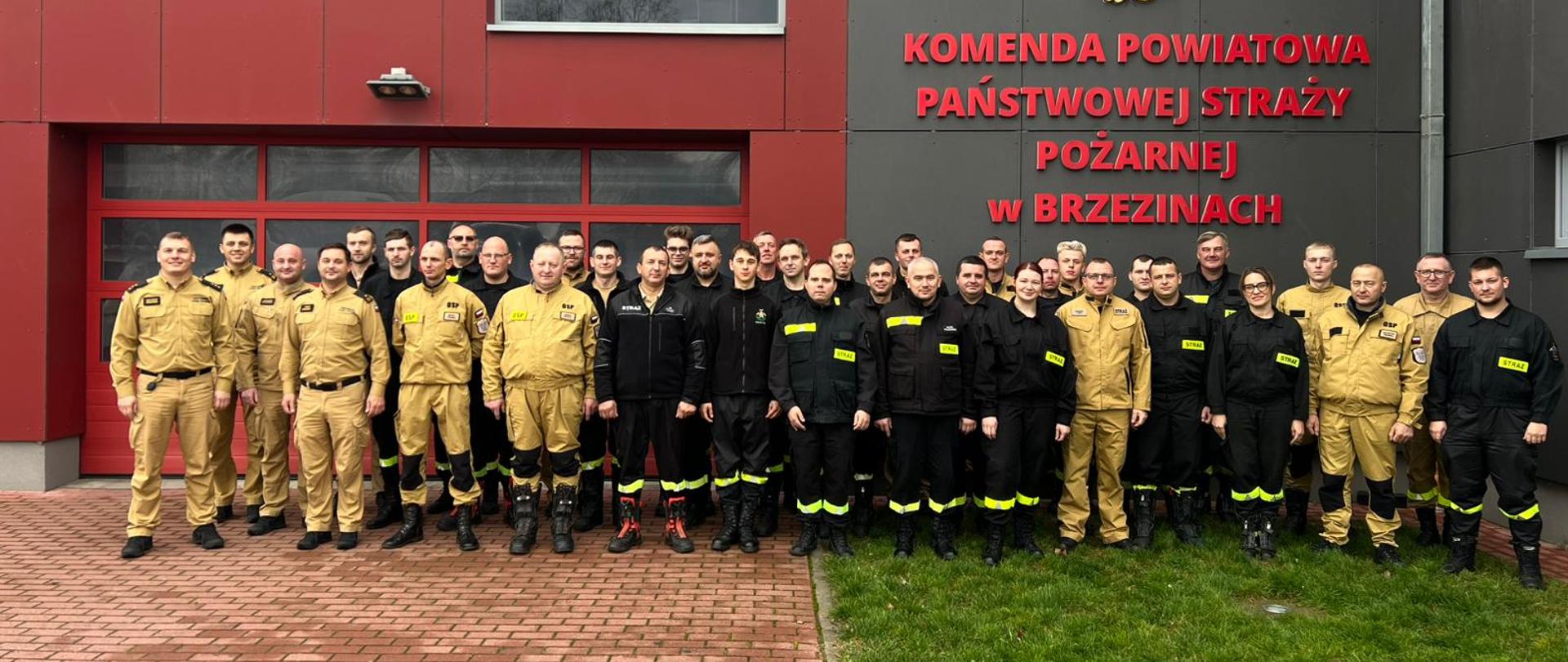 Egzamin końcowy szkolenia OSP, grupa uczestników szkolenia z Zastępcą Komendanta Powiatowego Państwowej Straży Pożarnej w Brzezinach młodszym brygadierem Jakubem Brzózką i członkami komisji egzaminacyjnej pozuje do zdjęcia na tle budynku strażnicy komendy. 