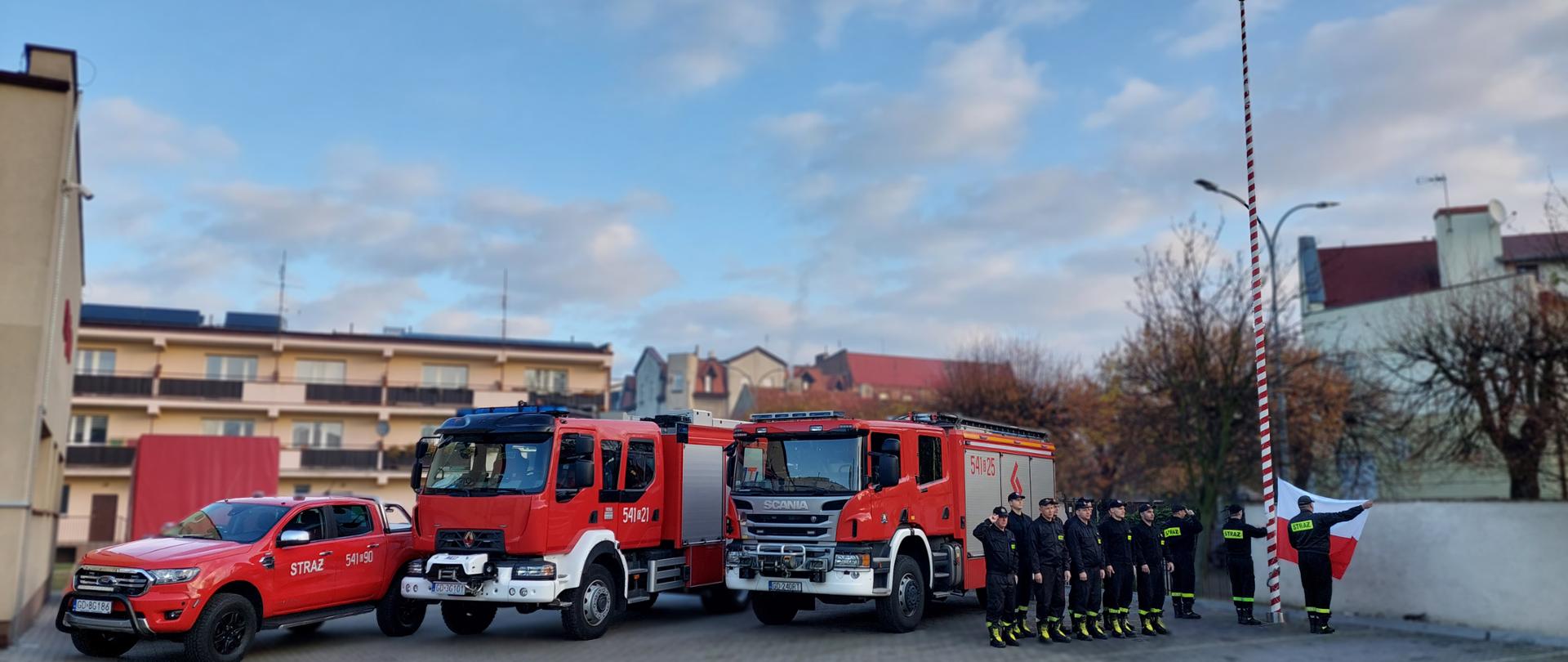 Na zdjęciu uroczysta zbiórka z okazji 105 rocznicy odzyskania przez Polskę niepodległości. Poczet flagowy zawiesza flagę Polski na maszt. Strażacy stoją w szyku. Za nimi ustawione są trzy samochody pożarnicze z Jednostki Ratowniczo-Gaśniczej w Tczewie.
