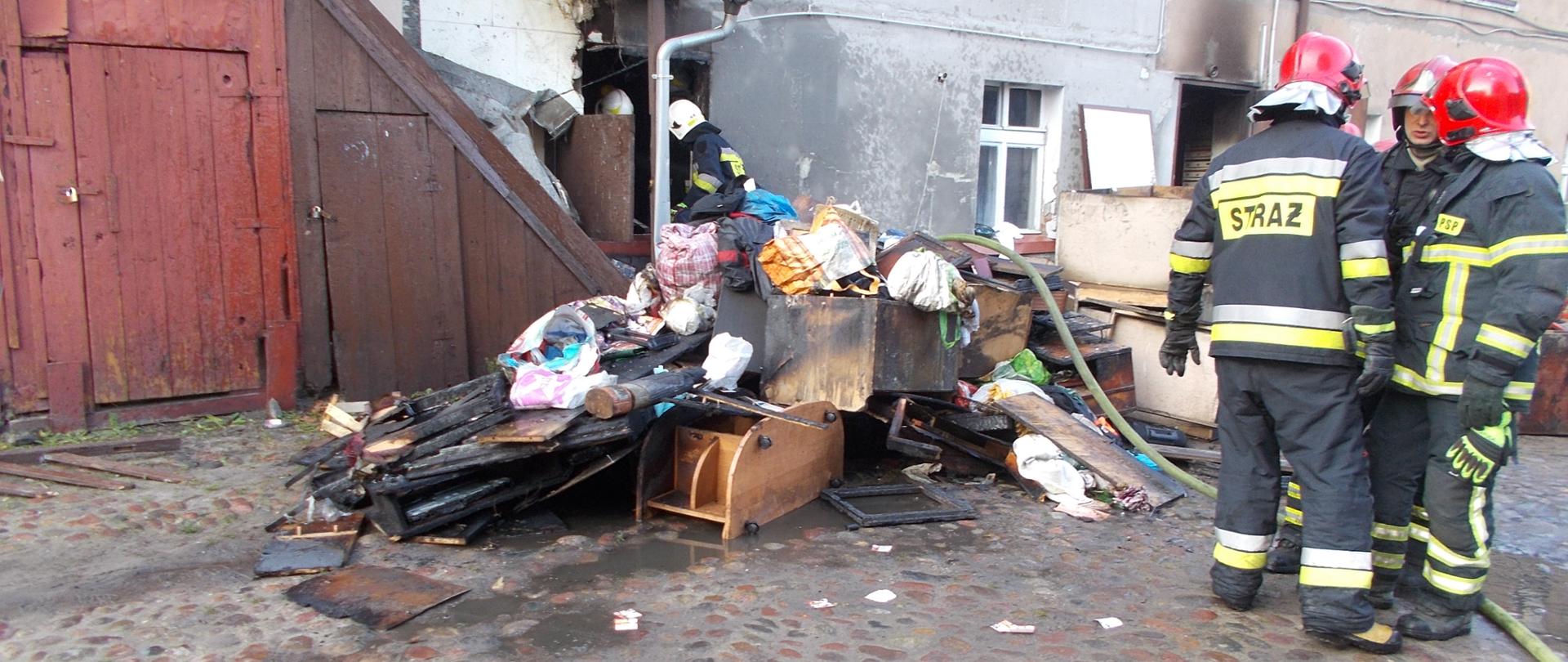 Strażacy w ciemnych mundurach i czerwonych hełmach stoją przed budynkiem przed którym zostały wyrzucone spalone i nadpalone materiały wyposażenia wnętrz: szafy, krzesła, półki. W tle budynek częściowo drewniany, częściowo murowany z szarą elewacją okopcony sadzą.